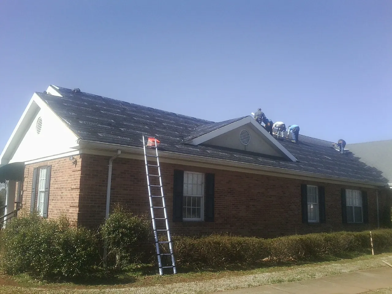 Crew tearing shingles off a brick ranch roof with a ladder set — Emergency Roof Repair work by our crew in Carle Place