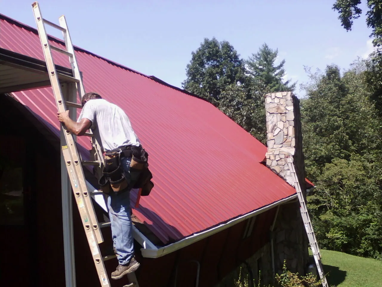 Roofer on a ladder beside a red metal roof and stone chimney — Soffit & Fascia Repair by Roofing Brothers in Carle Place