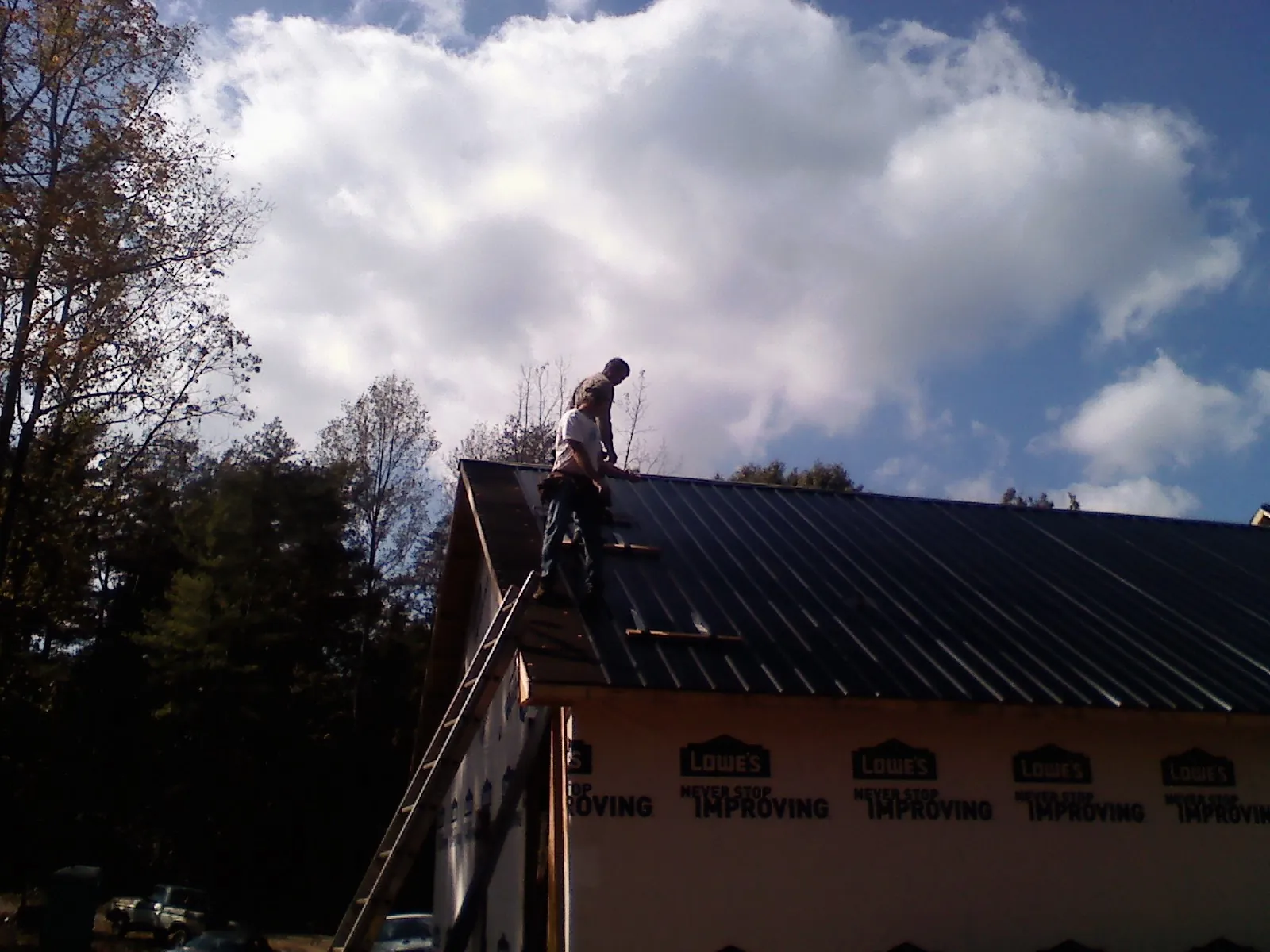 Roofer fastening dark metal panels high against a cloudy sky, from a Roof Inspection job in Carle Place